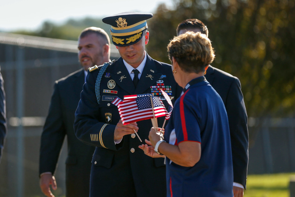 Donna Flinders, United Service Organizations volunteer, hands flags to those attending the funeral service for Army Master Sgt. Corey Hood, Saturday, Aug, 22, 2015, in West Chester, Ohio. Hood, 32, a member of the Army's Golden Knights parachute team, died Aug. 16 following a performance at the Chicago Air and Water Show.  Parachuters fly over Lakota West's football field Saturday, Aug. 21, 2015,  during the funeral service in West Chester, Ohio, for Army Master Sgt. Corey Hood. (Madison Schmidt/The Cincinnati Enquirer via AP)