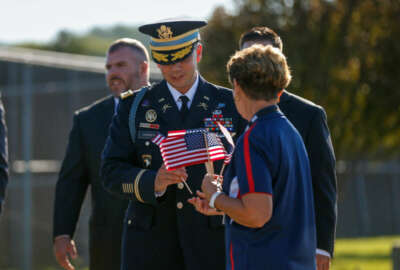 Donna Flinders, United Service Organizations volunteer, hands flags to those attending the funeral service for Army Master Sgt. Corey Hood, Saturday, Aug, 22, 2015, in West Chester, Ohio. Hood, 32, a member of the Army's Golden Knights parachute team, died Aug. 16 following a performance at the Chicago Air and Water Show.  Parachuters fly over Lakota West's football field Saturday, Aug. 21, 2015,  during the funeral service in West Chester, Ohio, for Army Master Sgt. Corey Hood. (Madison Schmidt/The Cincinnati Enquirer via AP)