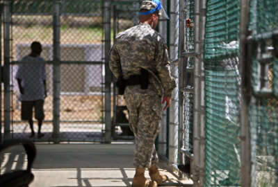 FILE - In this May 31, 2009 pool-file photo reviewed by the U.S. military and shot through a window, a guard wearing a protective face mask speaks with a detainee through a fence as another paces inside the exercise yard at Camp five detention facility on Guantanamo Bay U.S. Naval Base in Cuba. The Obama Administration’s struggling crusade to close the U.S. detention center at Guantanamo Bay, Cuba, is mired in state and federal politics, and frustrated White House and Pentagon officials are blaming each other for the slow progress releasing approved detainees and finding a new prison to house the remainder. (AP Photo/Brennan Linsley, Pool, File)