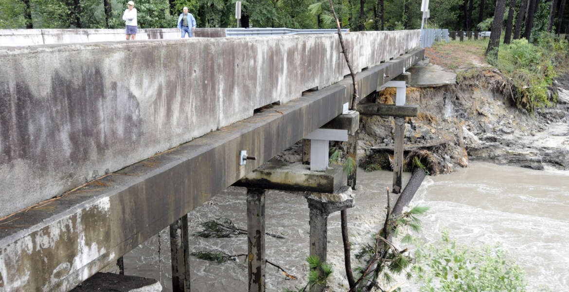 FILE- In this Monday, Oct. 5, 2015 file photo, Residents of a lakeside neighborhood walk across Overcreek Bridge by the remains of a failed dam in Columbia, S.C. Officials say the structure beneath a road collapsed Monday afternoon following days of heavy rain, nearly emptying a lake in a few minutes. No one was injured. South Carolina had problems with crumbling roads and bridges and old drinking water systems and dams long before the historic floods of the past week. Now the state faces what will likely be hundreds of millions if not billions of dollars of bills to fix washed out roads and bridges and destroyed dams. (AP Photo/Jay Reeves, File)