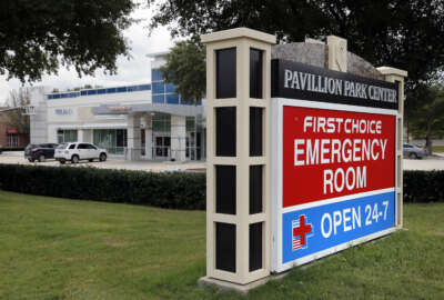 A sign by a nearby road intersection advertises a First Choice Emergency Room, rear, Thursday, Aug. 18, 2016, in Richardson, Texas. Freestanding emergency centers have sprouted in recent years across the suburban landscape, taking root in affluent neighborhoods and directly challenging nearby medical clinics and hospitals.  (AP Photo/Tony Gutierrez)