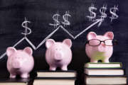 Three pink piggy banks standing on books next to a blackboard with simple savings progress chart.  Sharp focus on the piggy banks.  Alternative version shown below: