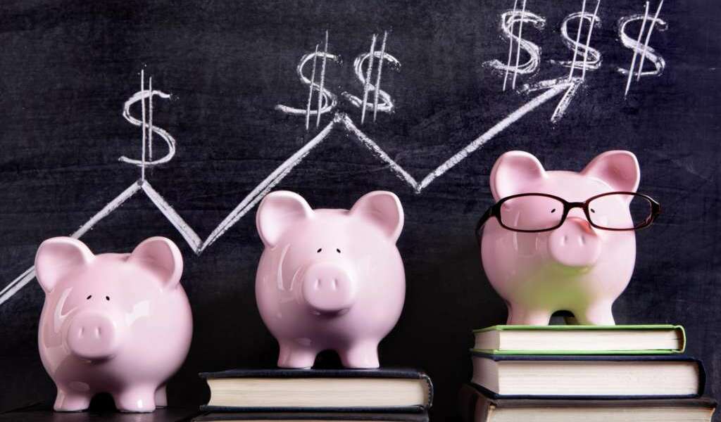 Three pink piggy banks standing on books next to a blackboard with simple savings progress chart.  Sharp focus on the piggy banks.  Alternative version shown below: