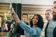 Young smiling black businesswoman at the wall with stickers communicating with her colleagues. Mixed team of entrepreneurs