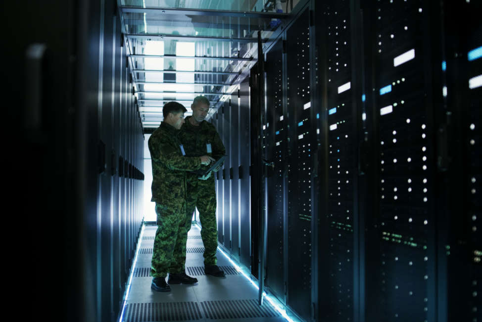 In Data Center Two Military Men Work with Open Server Rack Cabinet. One Holds Military Edition Laptop.