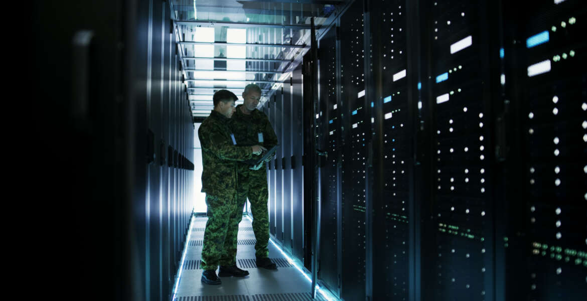 In Data Center Two Military Men Work with Open Server Rack Cabinet. One Holds Military Edition Laptop.