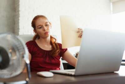 Portrait of young redhead woman working with computer laptop in office at summer during heatwave. The temperature is hot and the hair conditioner is broken. The girl sweats and feels exhausted