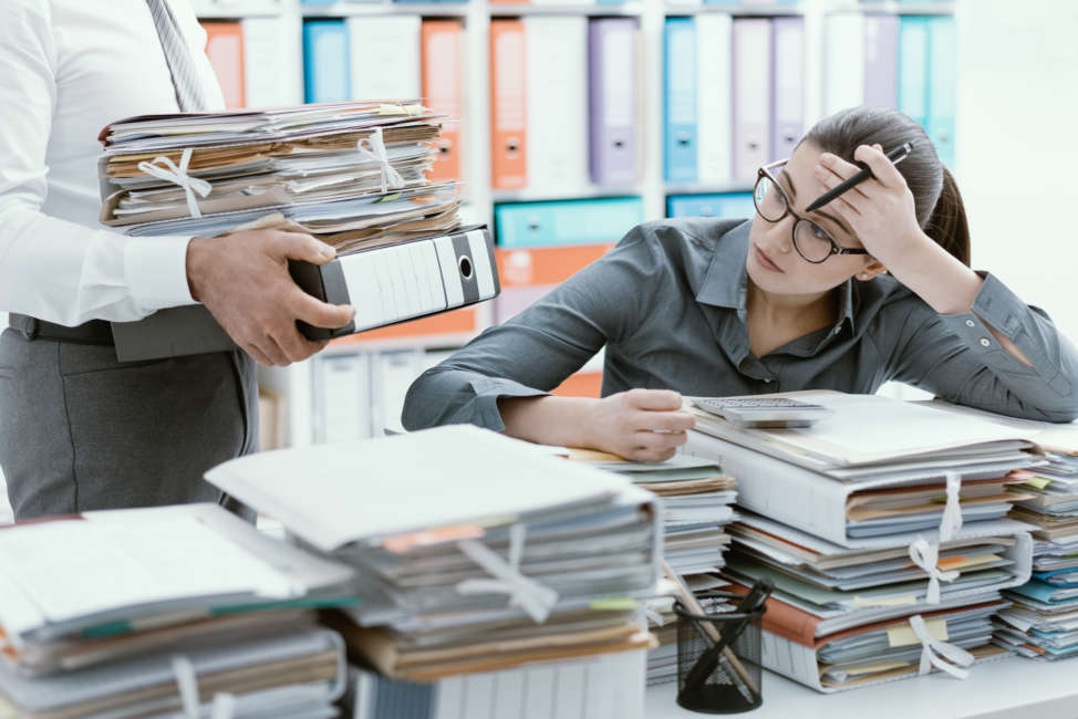 Young stressed secretary in the office overwhelmed by work and desk full of files, her boss is bringing more paperwork to her