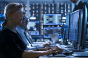 Female Government Employee Works in a Monitoring Room. In The Background Supervisor Holds Briefing. Possibly Government Agency Conducts Investigation.