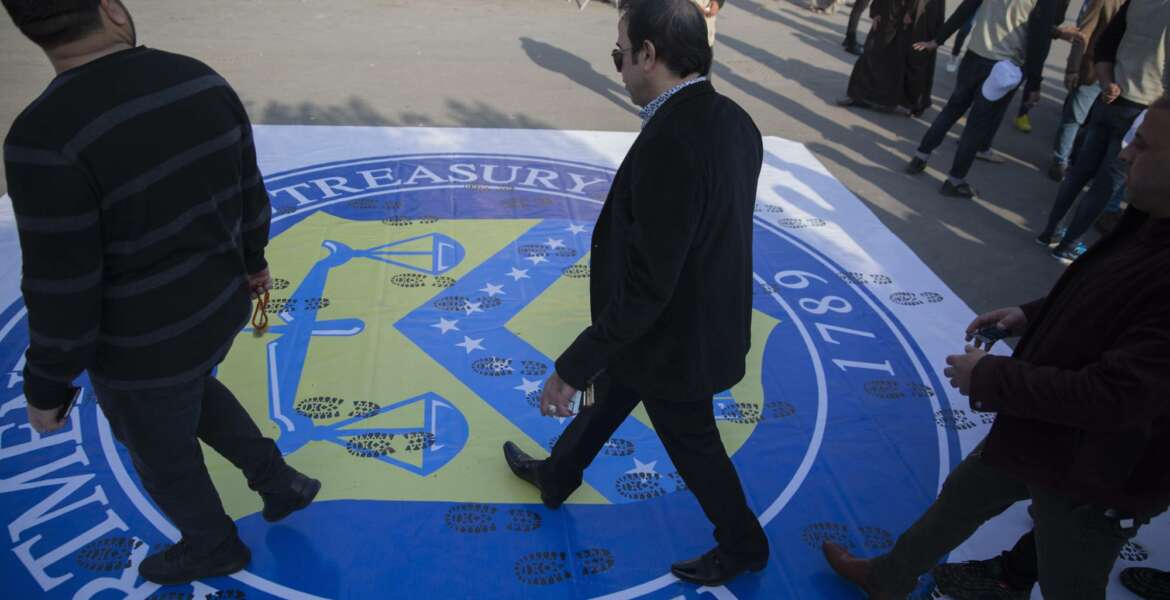Iraqi protesters walk over a big poster with the logo of the US Department Of The Treasury, that was spread on the street by the organizers to show disrespect, during a rally for the Shiite group Asaib Ahl al-Haq, in Baghdad, Iraq, Saturday, Dec. 14, 2019.   (AP Photo/Nasser Nasser)