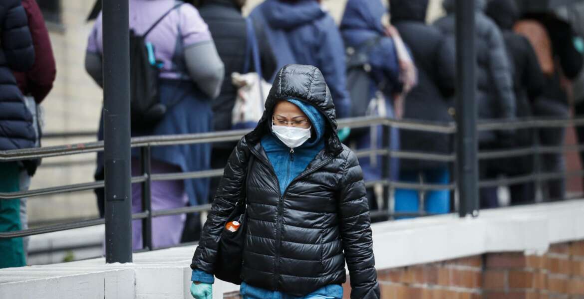 A medical worker wearing a single protective glove and a face mask walks past a line of workers and visitors waiting to be tested for COVID-19, the disease caused by the new coronavirus, at the main entrance to the Department of Veterans Affairs Medical Center, Monday, March 23, 2020, in New York.  (AP Photo/John Minchillo)