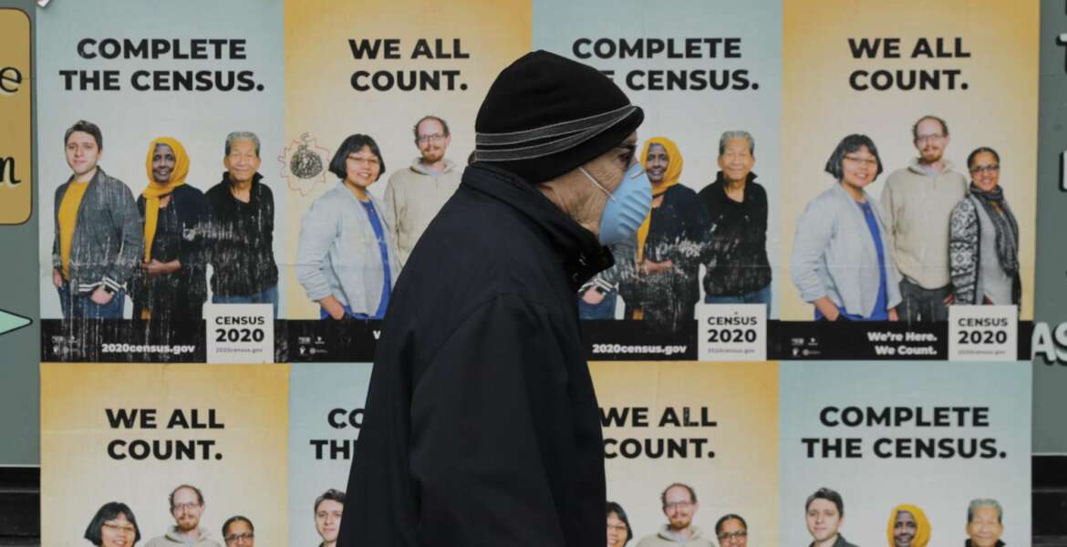 A man wearing a mask walks past posters encouraging participation in the 2020 Census, Wednesday, April 1, 2020, in Seattle's Capitol Hill neighborhood. Wednesday is Census Day, the date used to reference where a person lives for the once-a-decade count, as the U.S. is almost paralyzed by the spread of the novel coronavirus, but census officials vowed the job would be completed by its year-end deadline. (AP Photo/Ted S. Warren)