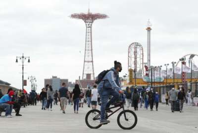 A young man wearing a protective face mask rides his bicycle along a fairly crowded Coney Island boardwalk during the current coronavirus outbreak, the afternoon of Sunday, May 24, 2020, in New York. No swimming was allowed and social distancing reminders were abundant on the beach as Memorial Day weekend kicked off the first weekend of summer. (AP Photo/Kathy Willens)