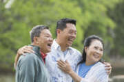 An Asian man standing in the park with his parents. The son is in the middle, with his arms around his parents shoulders. They are watching something, looking away from the camera, laughing