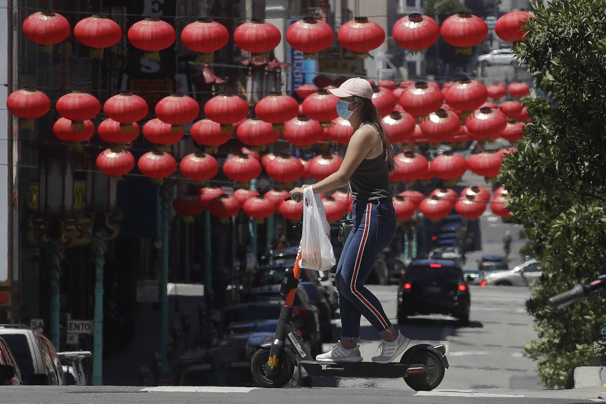 FILE - In this July 11, 2020, file photo, a woman wears a face mask while riding a scooter in front lanterns hanging in Chinatown during the coronavirus outbreak in San Francisco. In San Francisco, a city that depends largely on tourism, a nearly $11 billion loss in tourism spending is projected in 2020 and 2021 as the thousands of people who normally flock their for conventions and vacations have disappeared, according to the city's tourism bureau. (AP Photo/Jeff Chiu, File)