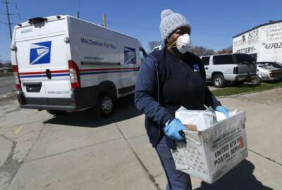 FILE - In this April 2, 2020 file photo, a United States Postal  Service worker makes a delivery with gloves and a mask in Warren, Mich. A group of states suing over service cuts at the U.S. Postal Service is asking a federal judge to immediately undo some of them, saying the integrity of the upcoming election is at stake.(AP Photo/Paul Sancya,File)