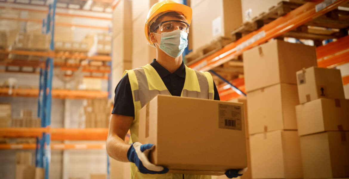 Portrait of Handsome Male Worker Wearing Medical Face Mask and Hard Hat Carries Cardboard Box Walks Through Retail Warehouse full of Shelves with Goods. Safety First Protective Workplace.