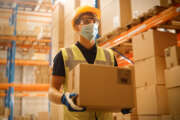 Portrait of Handsome Male Worker Wearing Medical Face Mask and Hard Hat Carries Cardboard Box Walks Through Retail Warehouse full of Shelves with Goods. Safety First Protective Workplace.