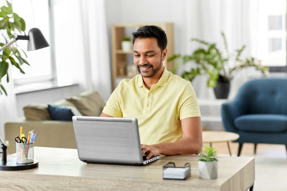 technology, remote job and lifestyle concept - happy smiling indian man with laptop computer working at home office